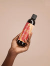 Hand hält Flasche As I Am Jamaican Black Castor Oil Leave-in Conditioner vor beigem Hintergrund